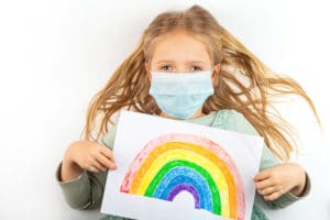photo of young girl wearing mask holding a crayon drawing of a rainbow